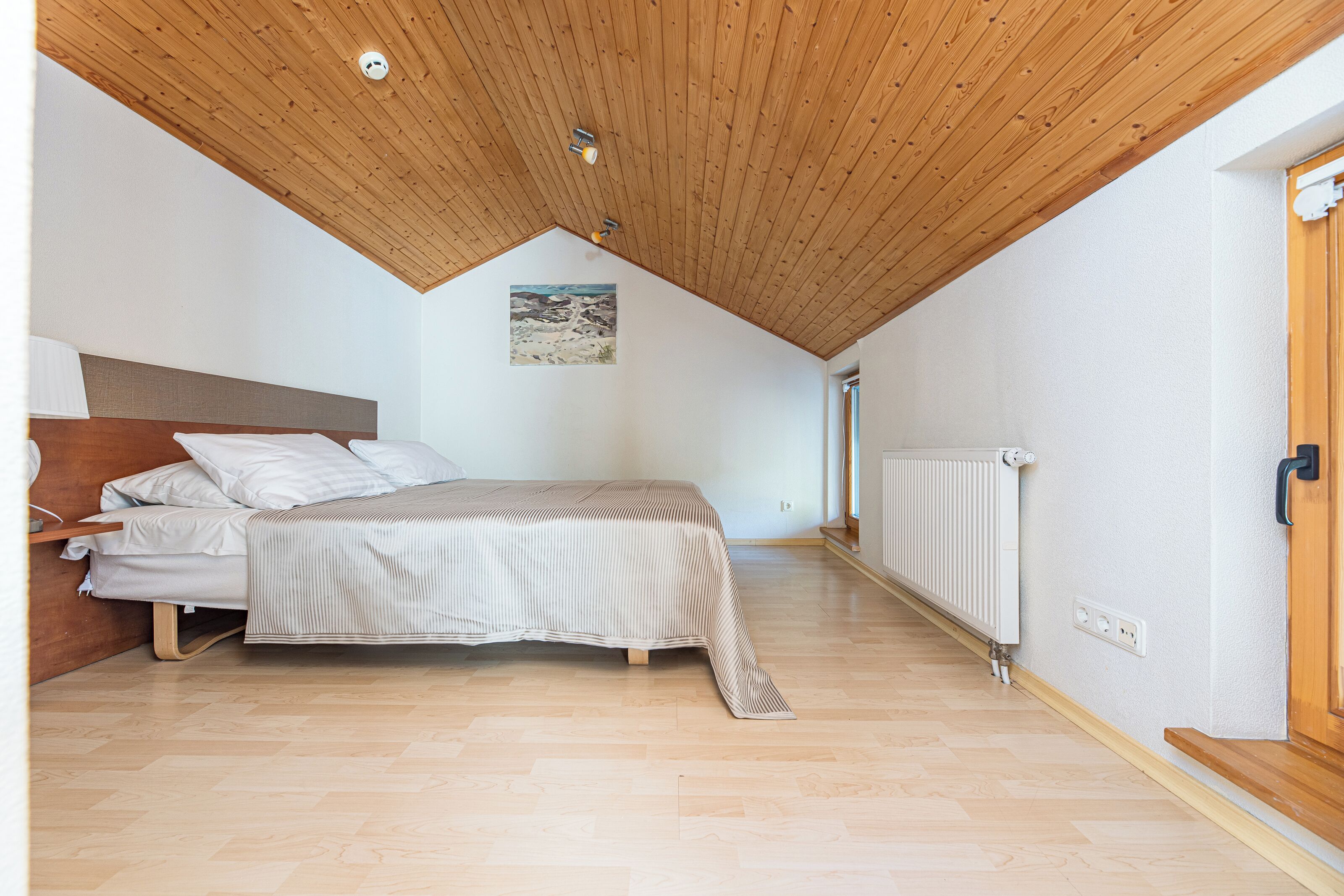 Un dormitorio con techo de madera, una cama, un cabecero, un radiador y una ventana.