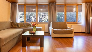 Ein Wohnzimmer mit Sofa, Sessel und einem Tisch mit Blumen. Große Fenster mit Vorhängen.
