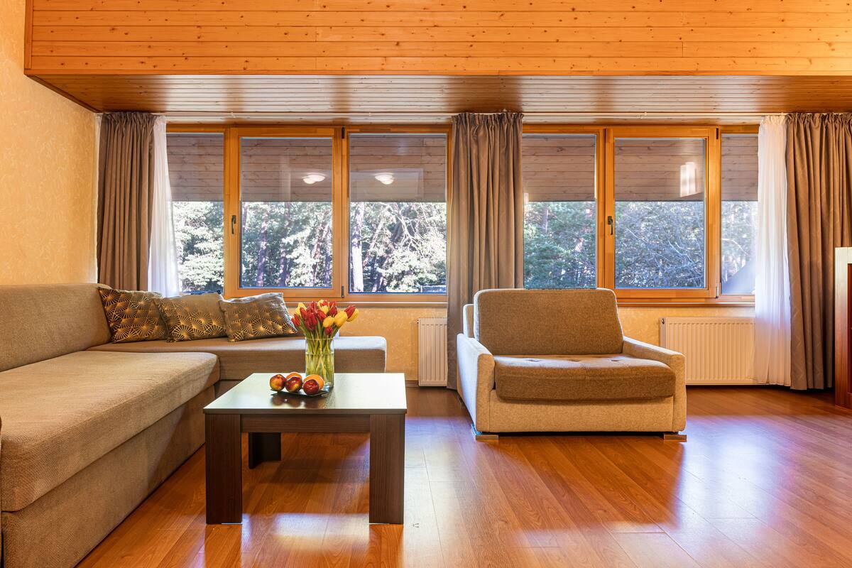 A living room with a sofa, armchair, and a table with flowers. There are large windows with curtains.