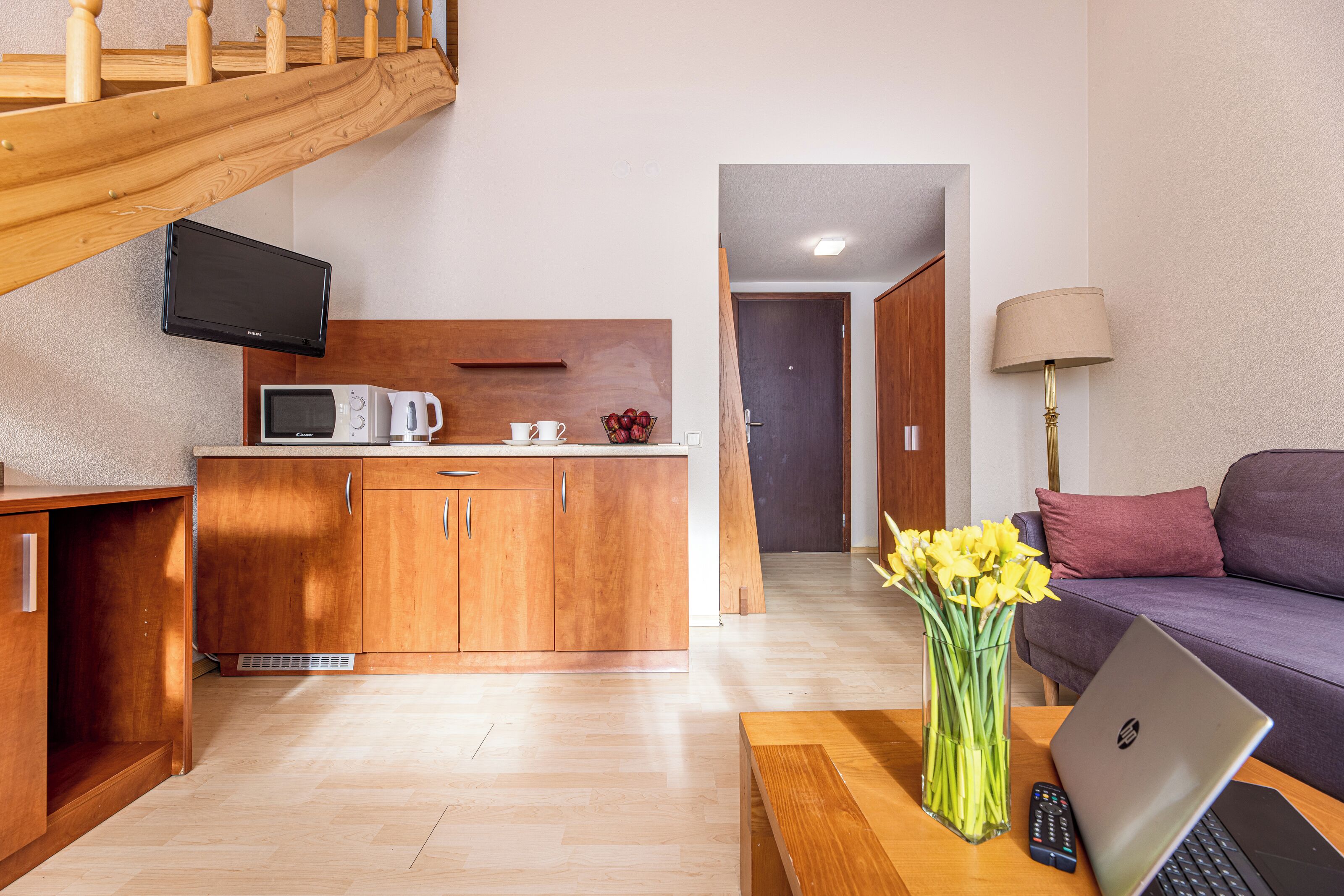 Una sala de estar moderna con una escalera de madera, una zona de cocina con microondas y un sofá con una computadora portátil y flores.