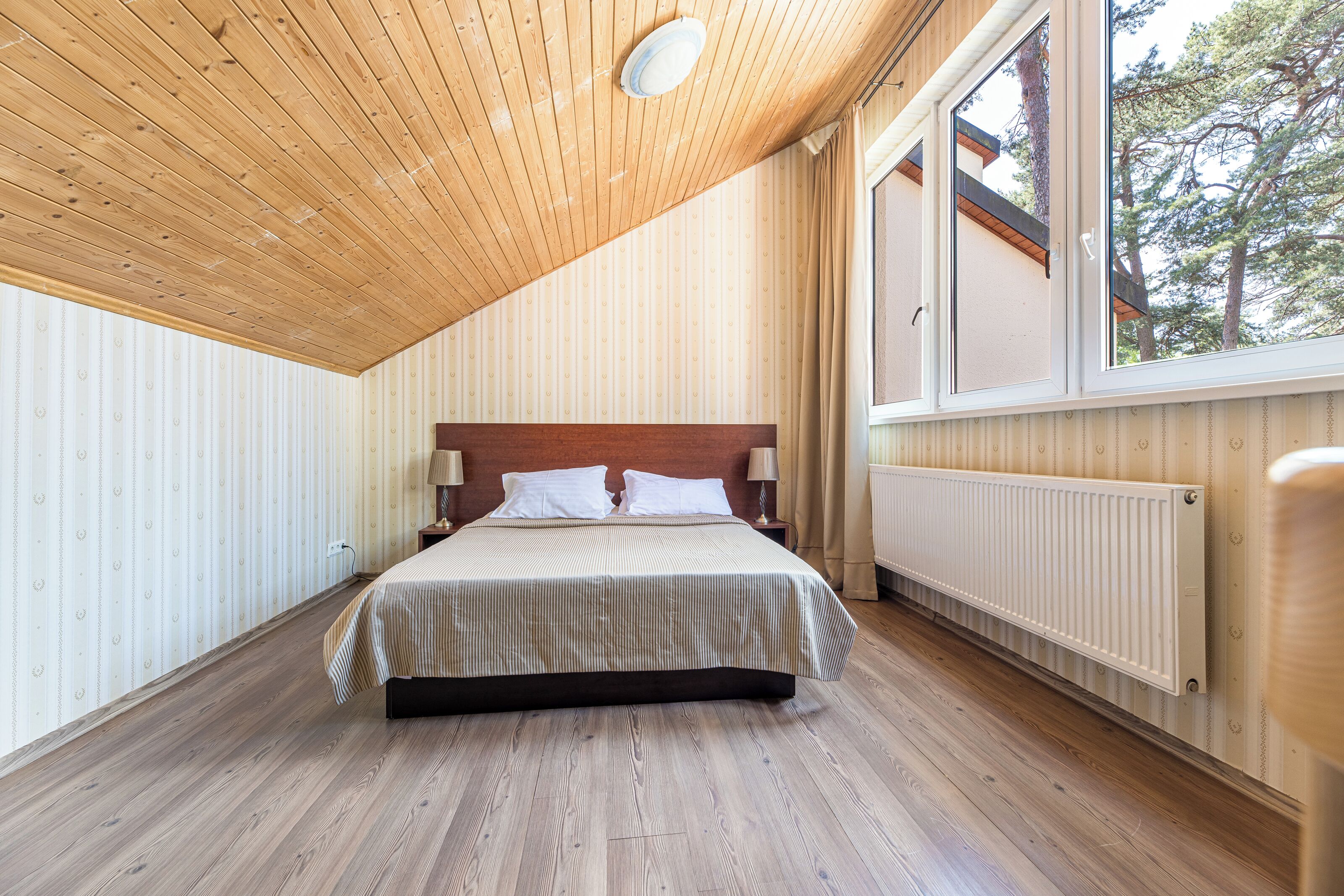 Un dormitorio con techo de madera, una cama grande con ropa de cama blanca, dos lámparas de noche y una ventana con vistas a árboles.