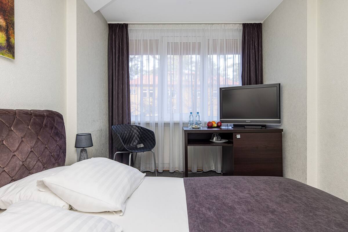 A hotel room with a bed, a desk with a television, a chair, and a window with curtains.