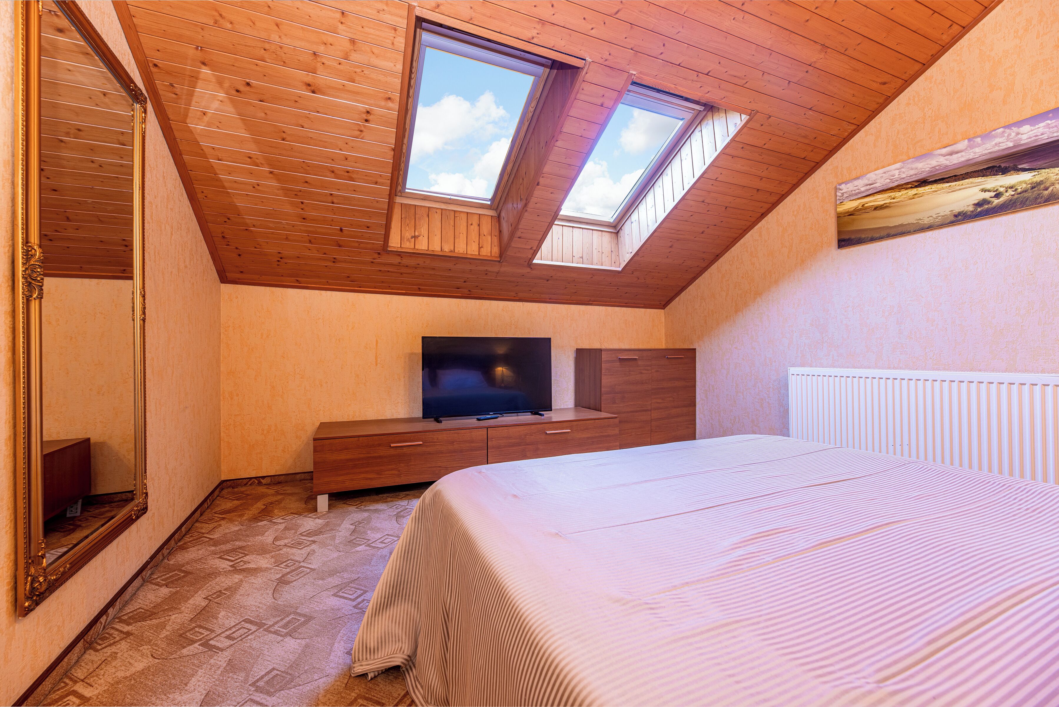 Un dormitorio con techo inclinado, tragaluces, cama, televisor y cómoda.