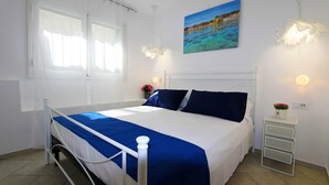 Un lit bien fait, recouvert d’une couvre-lit bleue et blanche, une table de chevet blanche ornée d’un vase de fleurs et, au mur, un tableau représentant un paysage côtier.