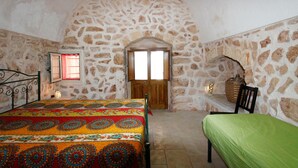 A bedroom with a stone wall, a bed with a colorful bedspread, a chair, a wooden door, and a small table with a lamp. - Trullo Saracino (Salve)