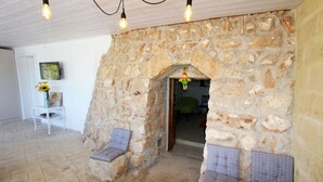 A stone-walled room with two cushioned seating areas, a door leading to another room, and a wall-mounted picture. - Trullo Saracino (Salve)