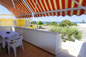Uma varanda com um toldo listrado, uma mesa com cadeiras e vista para o mar e edifícios circundantes.