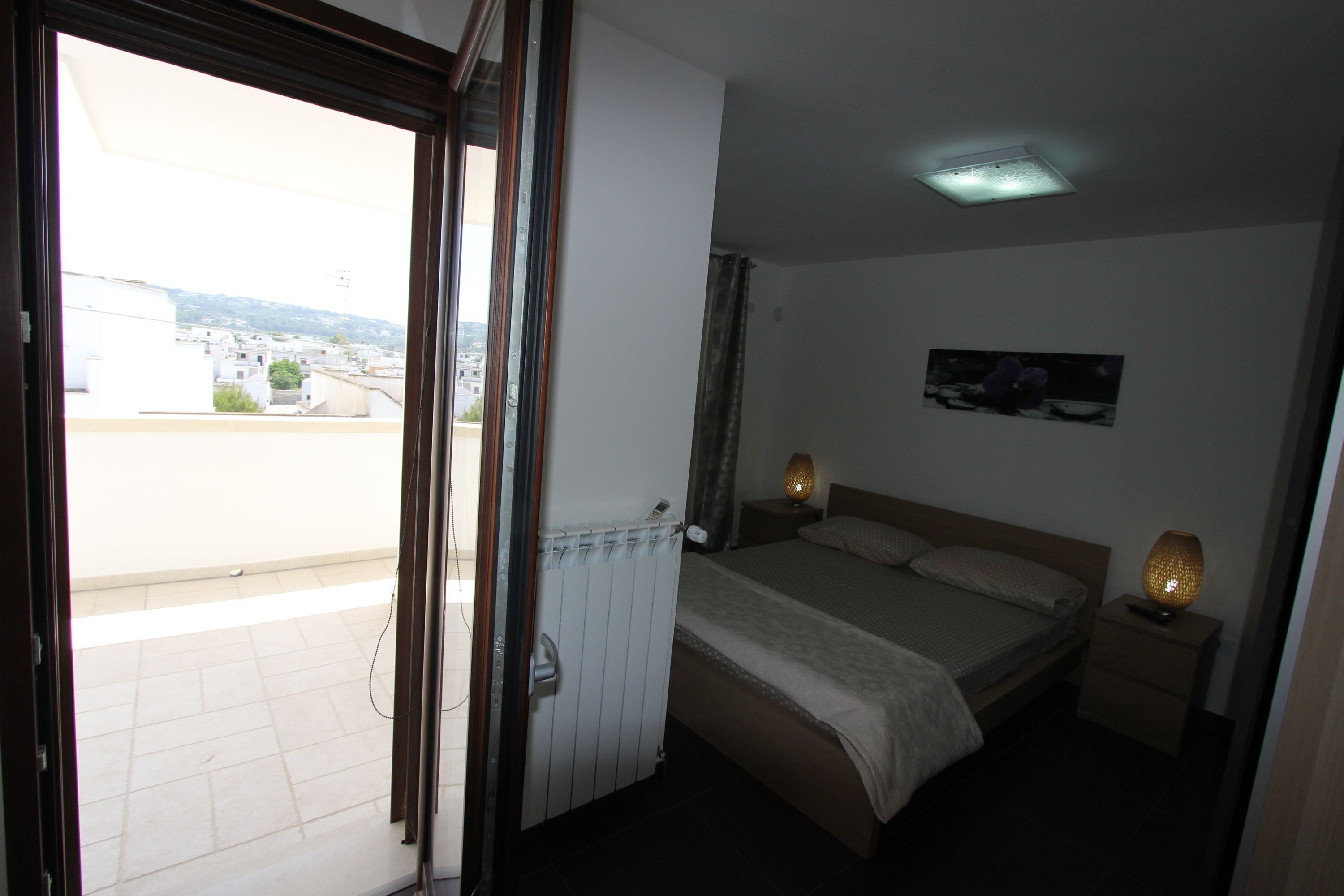 Un dormitorio con cama, lámparas de noche, una mesita de noche, un radiador, una ventana y vistas a edificios.
