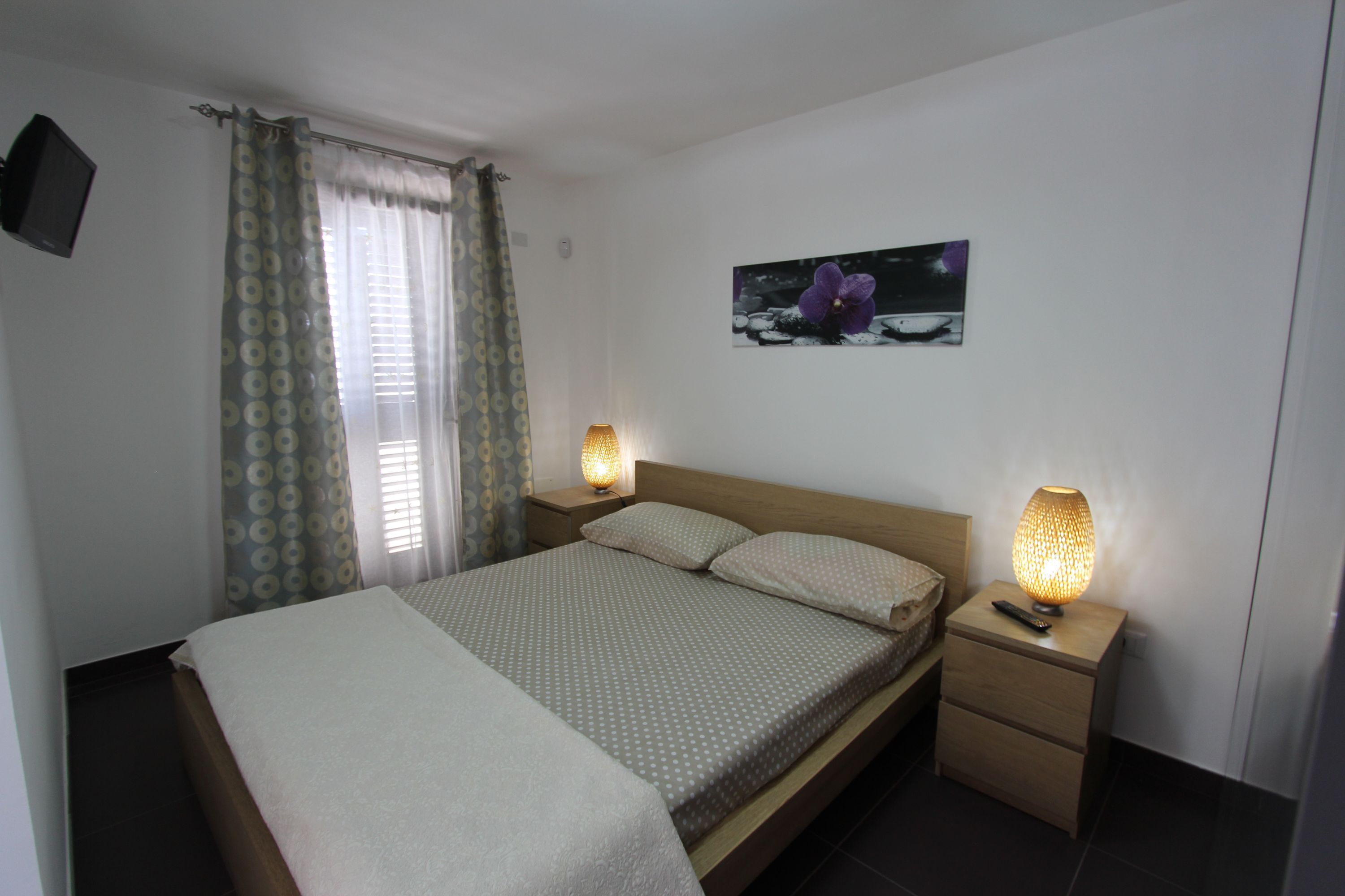 A bedroom with a bed, two bedside tables with lamps, a wall-mounted TV, and a framed picture.