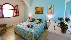 A bedroom with a bed, a chair, a nightstand, a bedside table, a lamp, a mirror, and a potted plant. - Villetta Mare 464 (Patu)