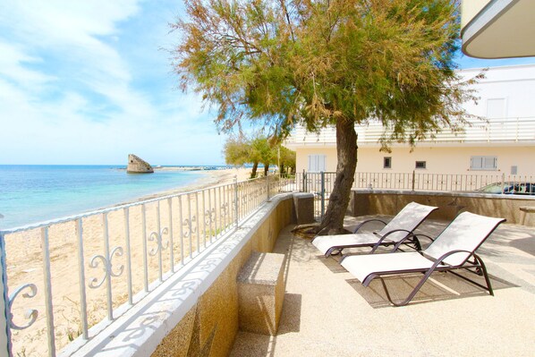A beachside balcony with two sun loungers, a tree, and a view of the sea and a rock formation. - Beach House in Puglia (Salve)