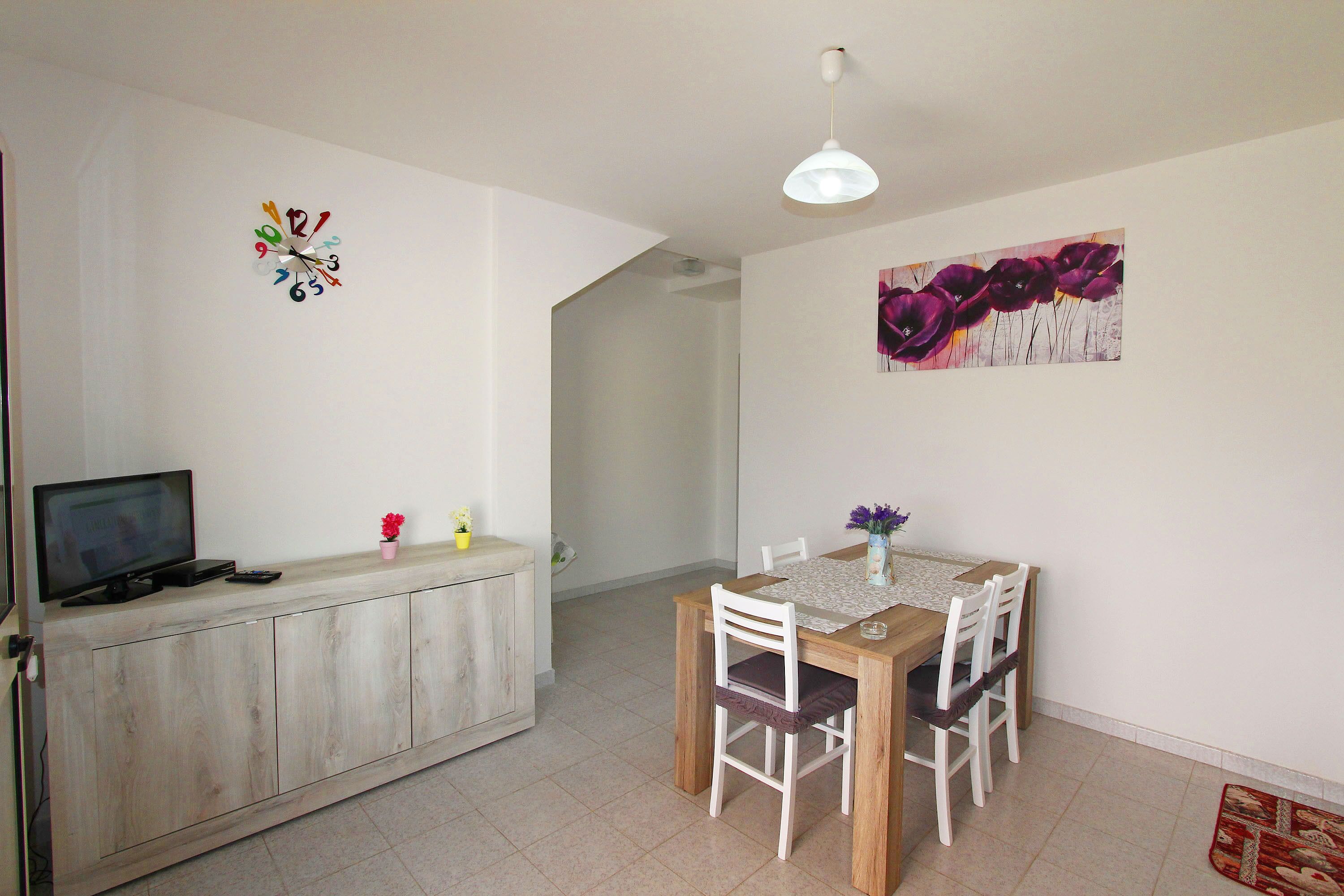 A dining area with a wooden table and chairs, a television on a cabinet, and a wall clock.