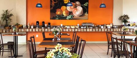 A restaurant with multiple tables and chairs, a fruit painting on the wall, and hanging blue pendant lights.