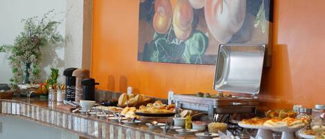Ein Buffet mit verschiedenen Speisen, Kaffeeautomaten und einem Gemälde mit Früchten an der Wand.