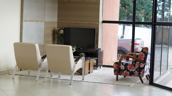 Una sala de estar moderna con dos sillones beige, un sofá estampado, una mesa de centro de vidrio y un televisor de pantalla plana sobre un mueble de madera.