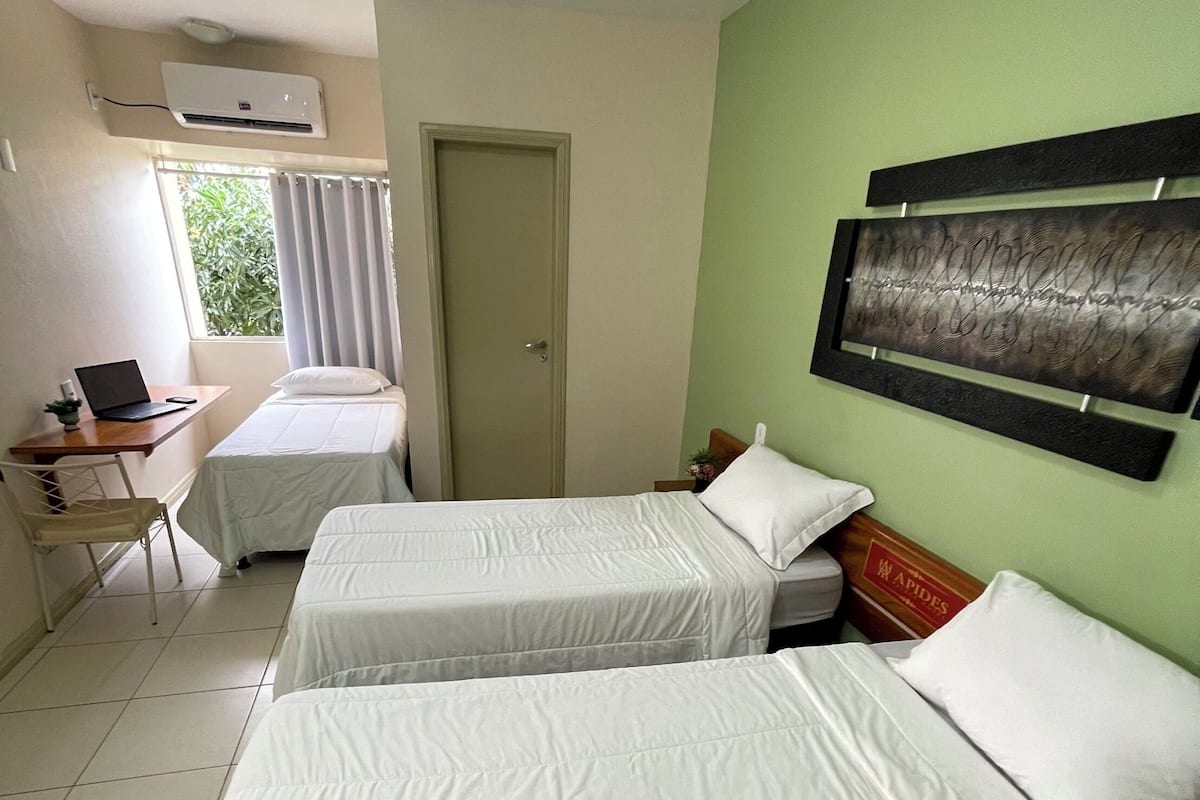 Ein Hotelzimmer mit zwei Betten, einem Schreibtisch mit einem Laptop, einem Fenster mit Vorhängen und einem an der Wand hängenden Kunstwerk.