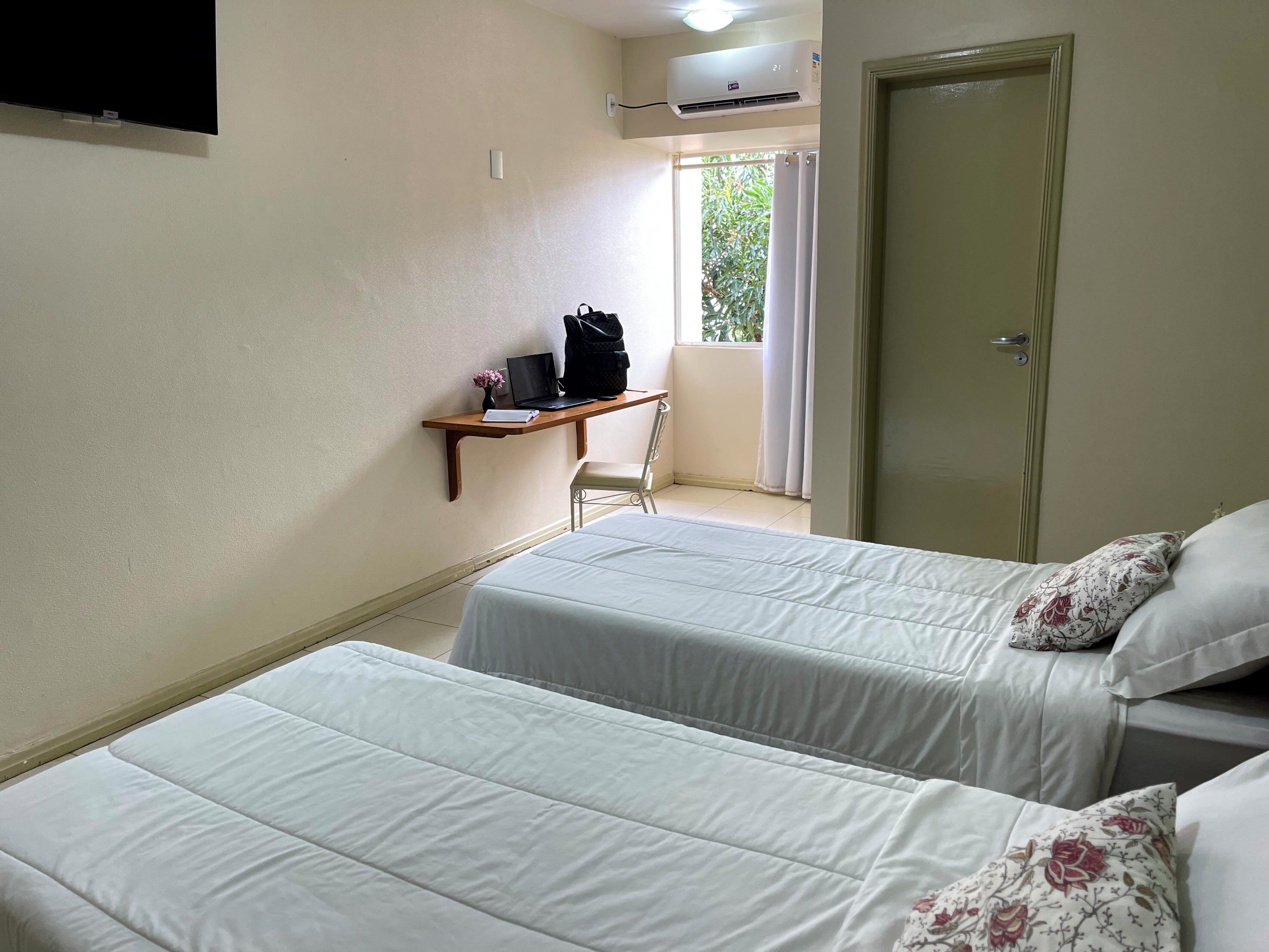 A room with two beds, a desk with a laptop, a chair, and a window with curtains.