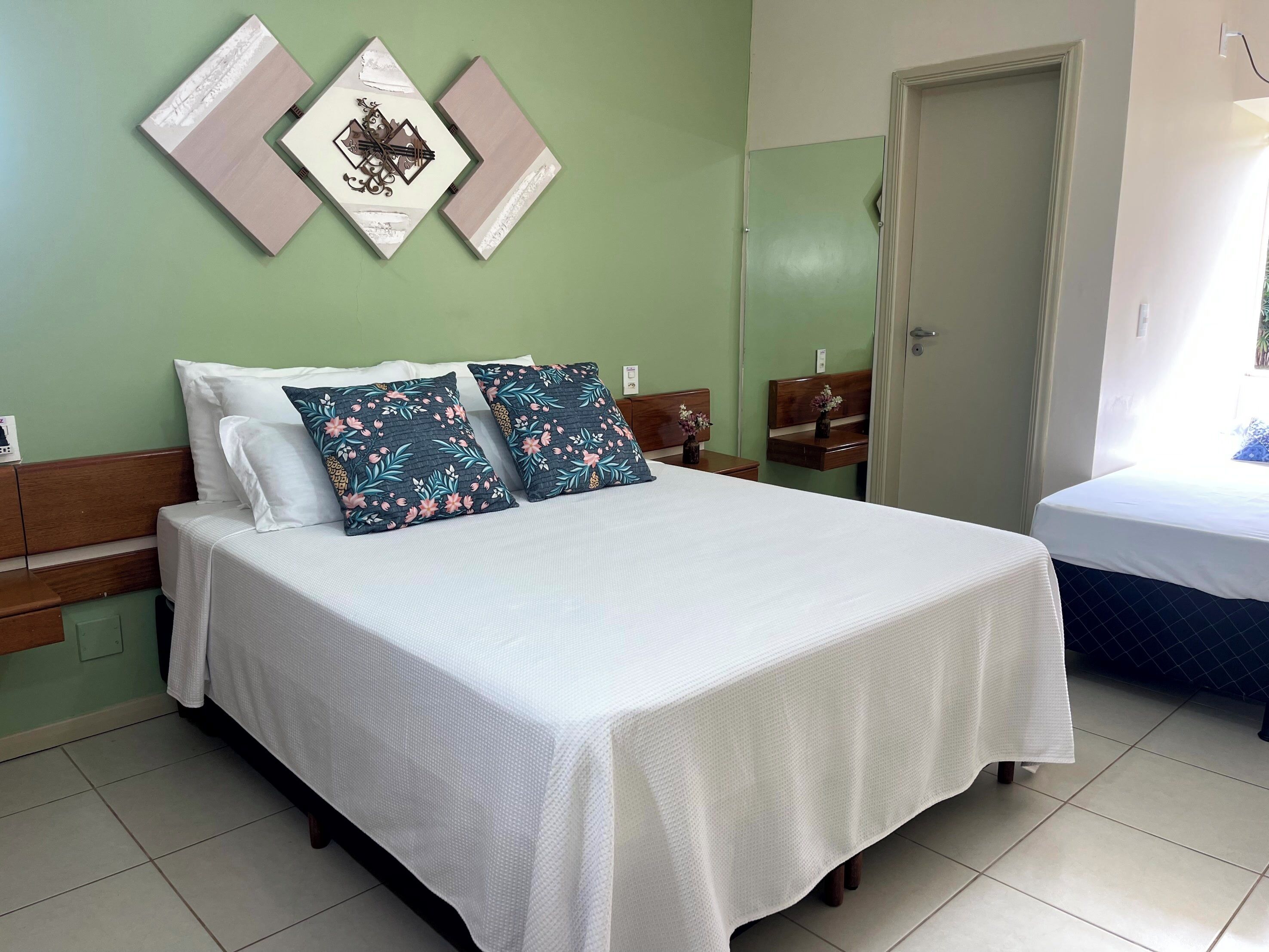 A neatly made bed with white bedding and two floral pillows, a wooden headboard, and a green wall with three framed pictures.