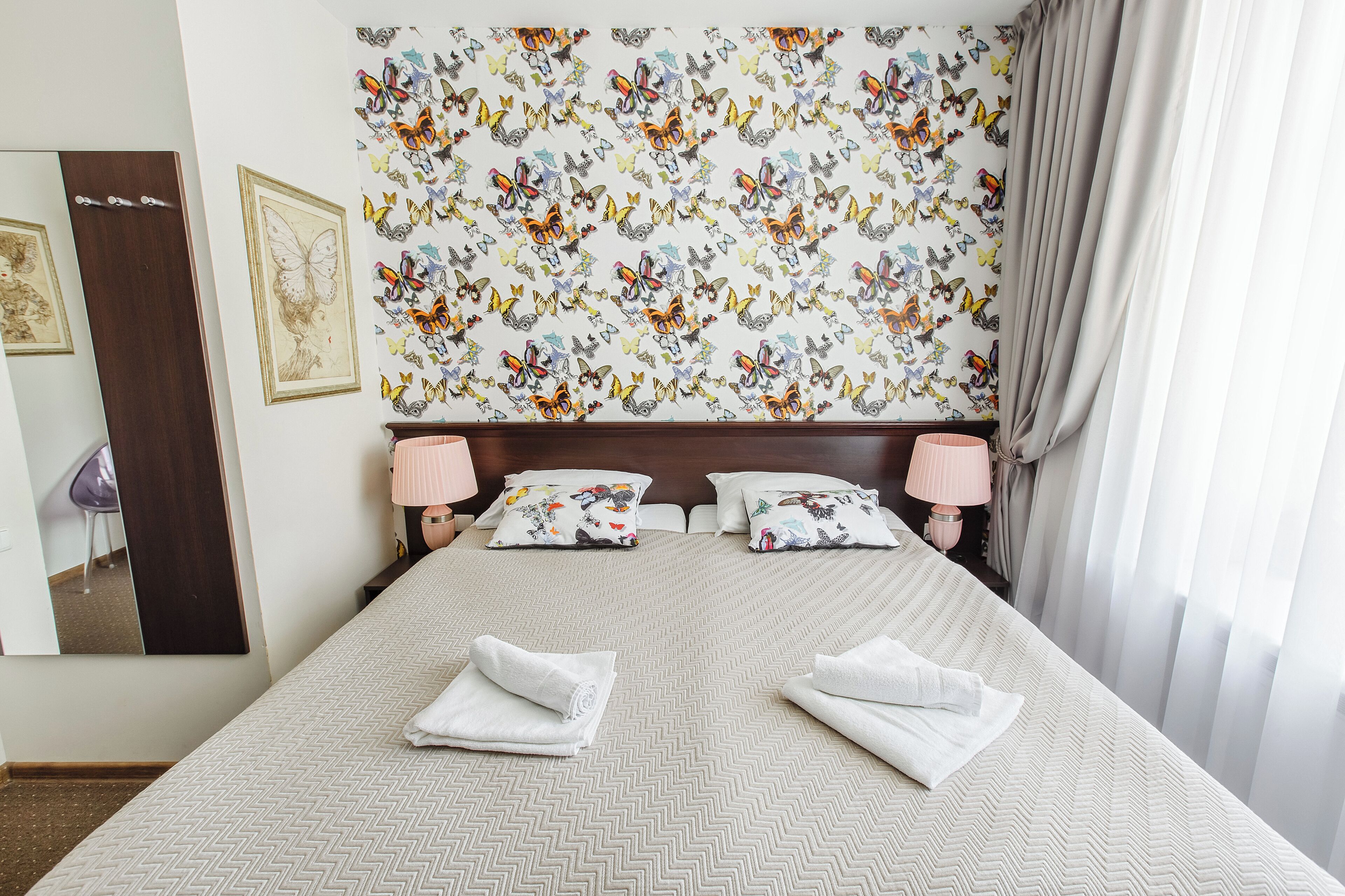 Un letto rifatto con cura, con una coperta a motivi e due cuscini decorati con un disegno di farfalla, e uno specchio appeso al muro.