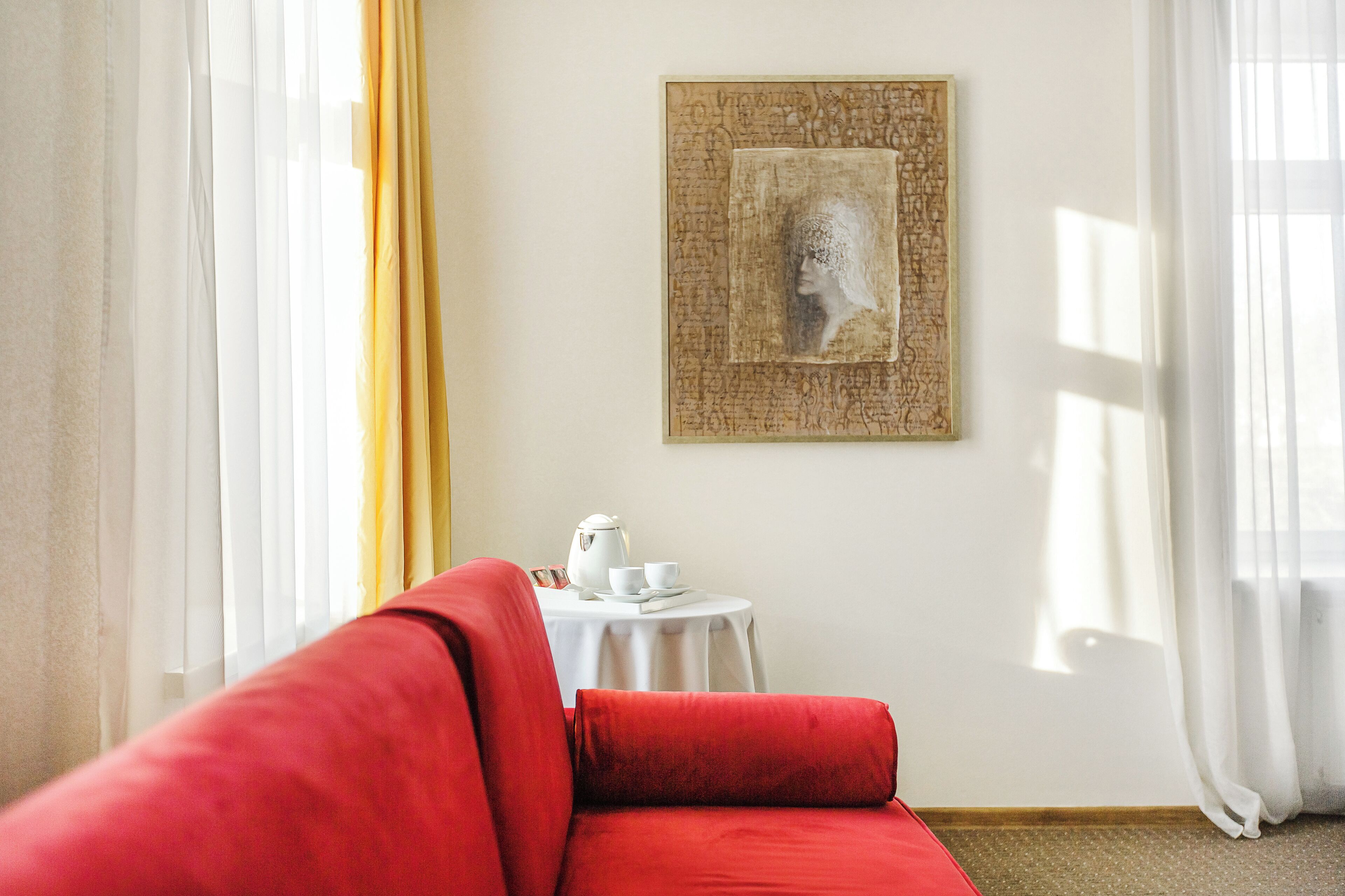 Una habitación con un sofá rojo, una mesita redonda con una tetera, tazas y platillos, y una obra enmarcada en la pared.