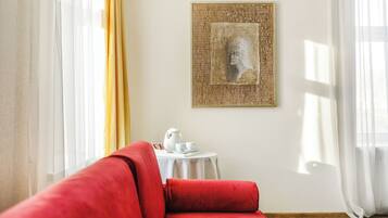 Una habitación con un sofá rojo, una mesita redonda con una tetera, tazas y platillos, y una obra enmarcada en la pared.