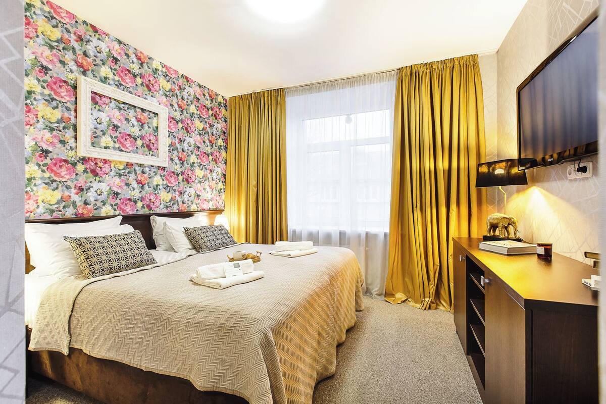 A hotel room with a floral wallpaper, a bed with a beige bedspread, a wooden dresser, and a television on the wall.