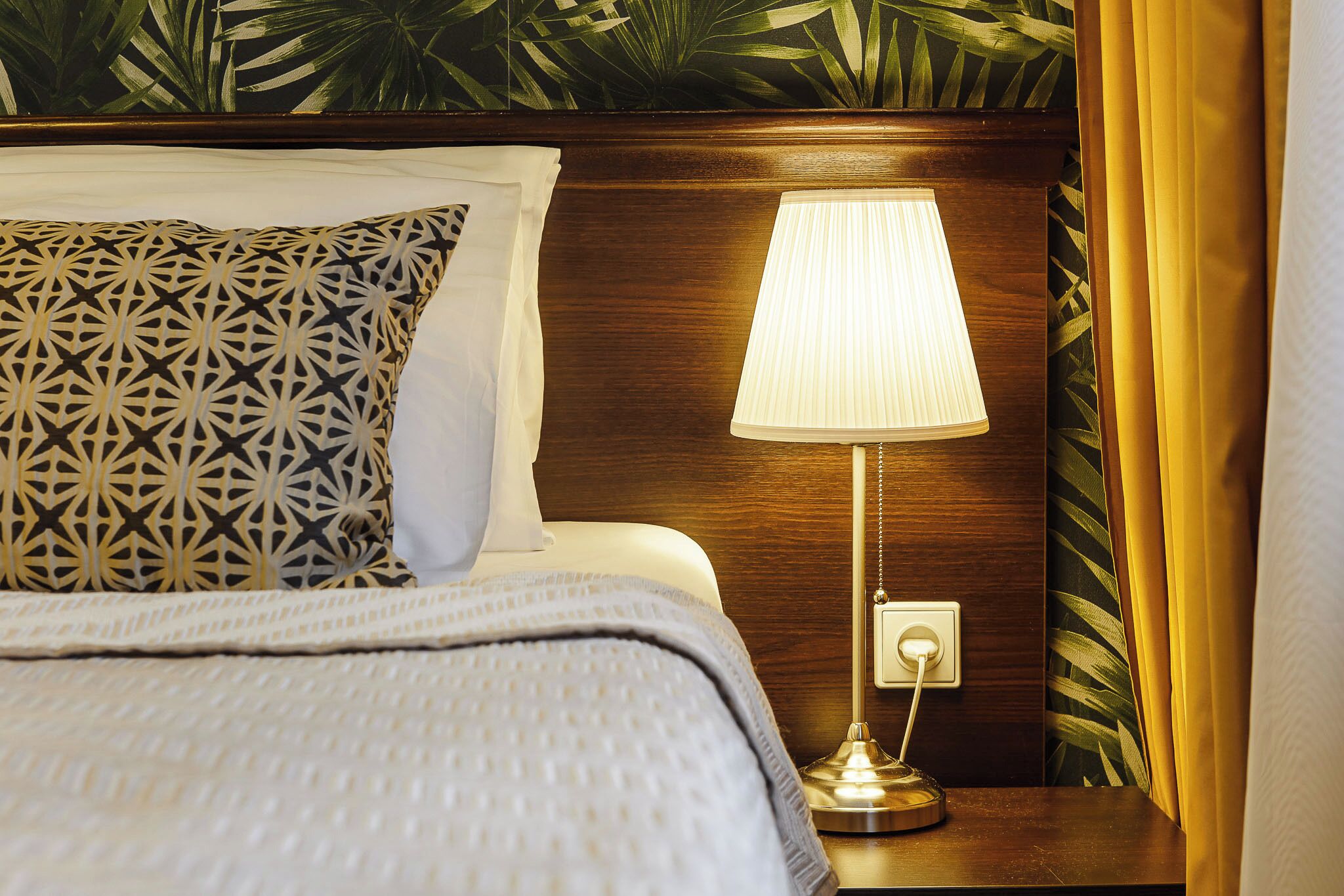 A neatly made bed with patterned pillows, a bedside lamp, and a wall with a leafy pattern.