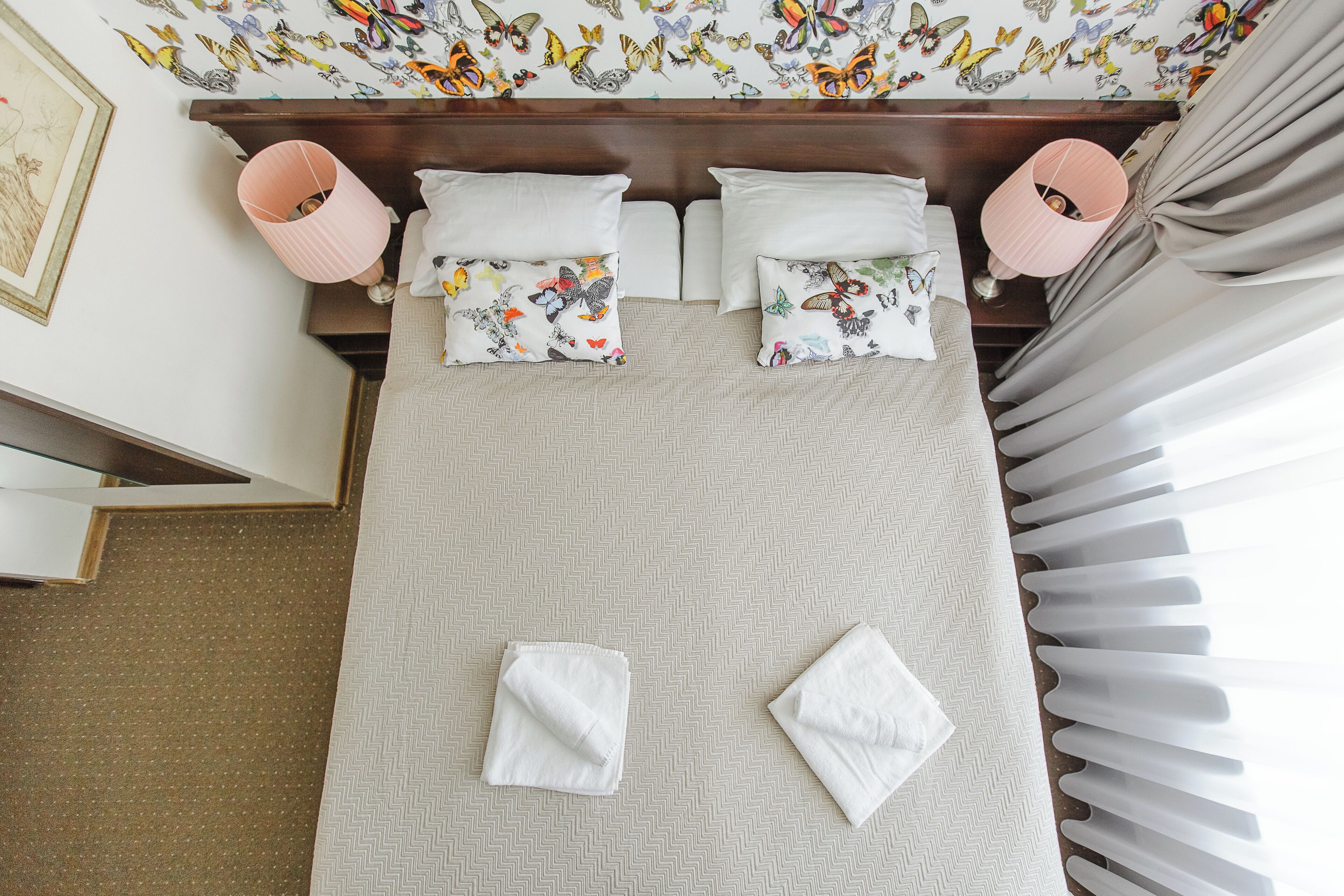 Una cama bien hecha con almohadones florales, un cabecero de madera y una pared decorada con adhesivos de mariposas.
