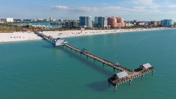 A pier extending into a clear blue sea with a beach and buildings in the background.