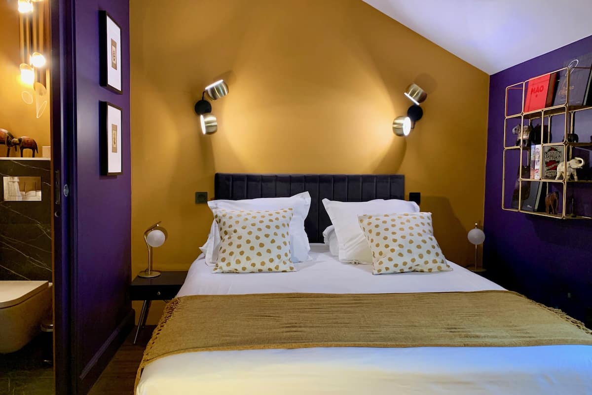 A hotel room with a bed, two bedside tables, a nightstand, a wall-mounted shelf with books, and a door with a mirror.