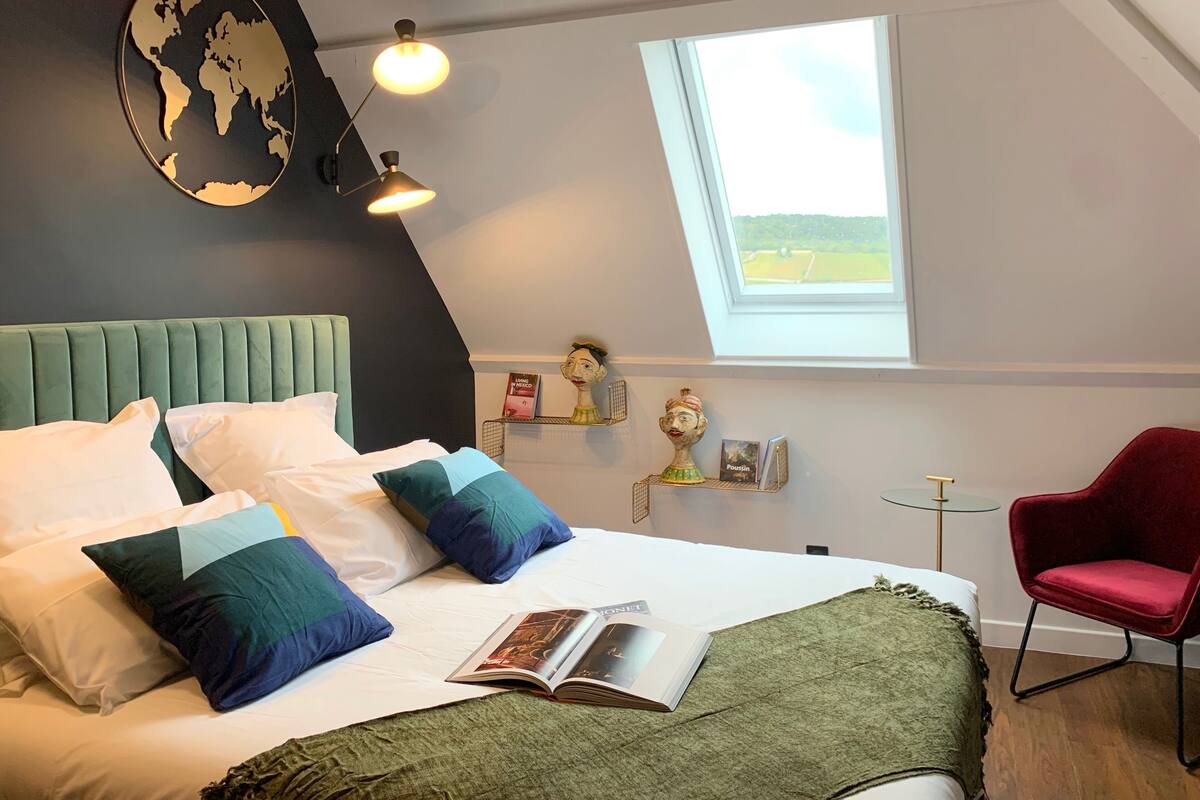 A bedroom with a bed, pillows, a book, a red chair, a small table, a globe wall decoration, and a window with a view.