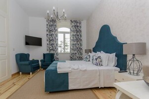 Habitación de hotel con cama, dos sillones, televisión, lámpara de araña y ventana con cortinas.