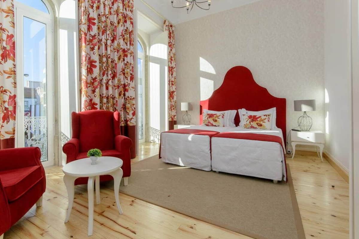 Ein Schlafzimmer mit rotem Kopfteil, einem Bett mit weißer Bettwäsche, einem roten Sessel, einem kleinen runden Tisch mit einer Pflanze und Blick auf die Stadt durch große Fenster.
