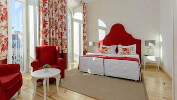 Quarto com cabeceira vermelha, cama com roupa de cama branca, poltrona vermelha, mesa redonda pequena com uma planta e vista da cidade através de grandes janelas.