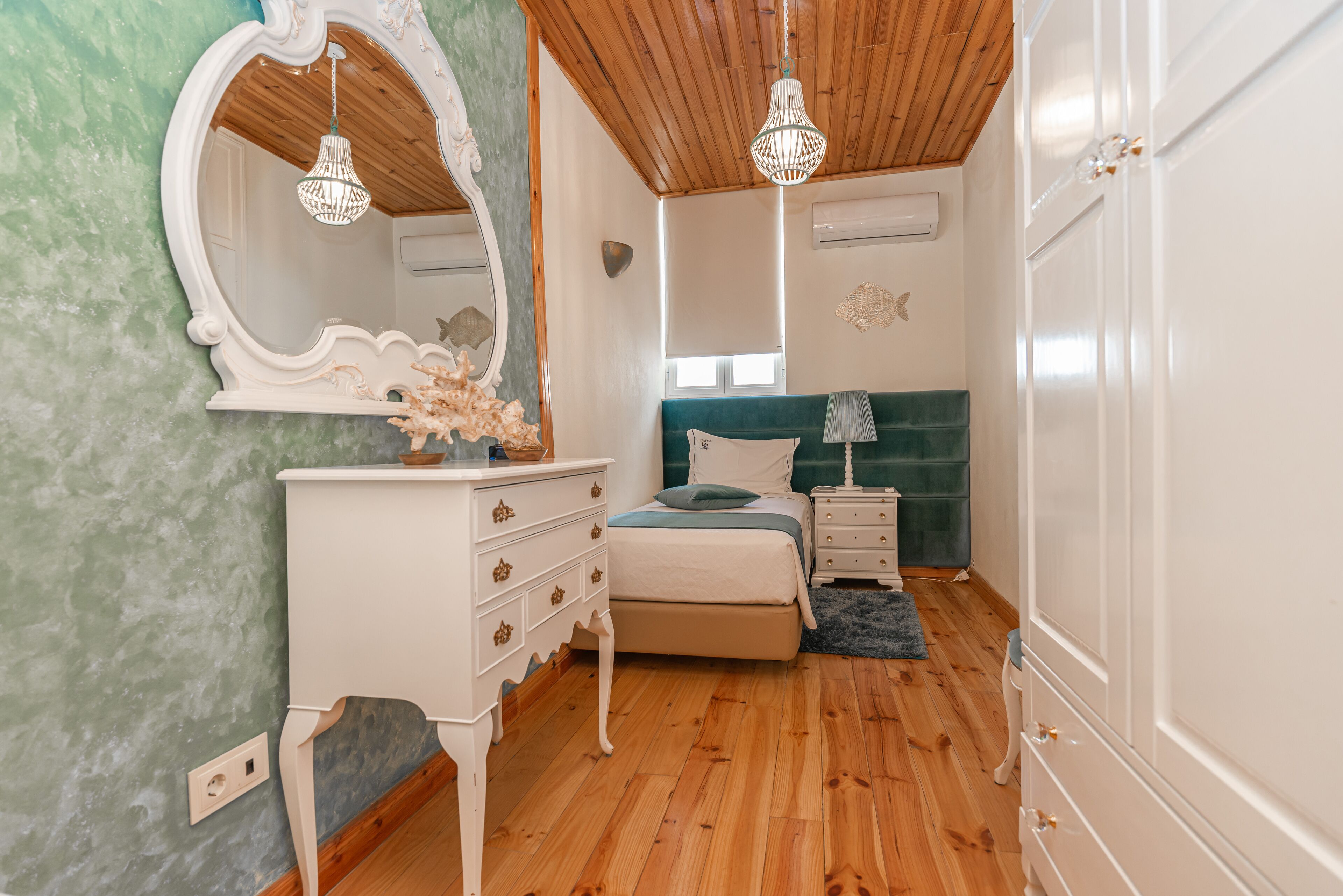 A bedroom with a wooden ceiling, a bed with a green headboard, a white dresser, a mirror, and two bedside tables.