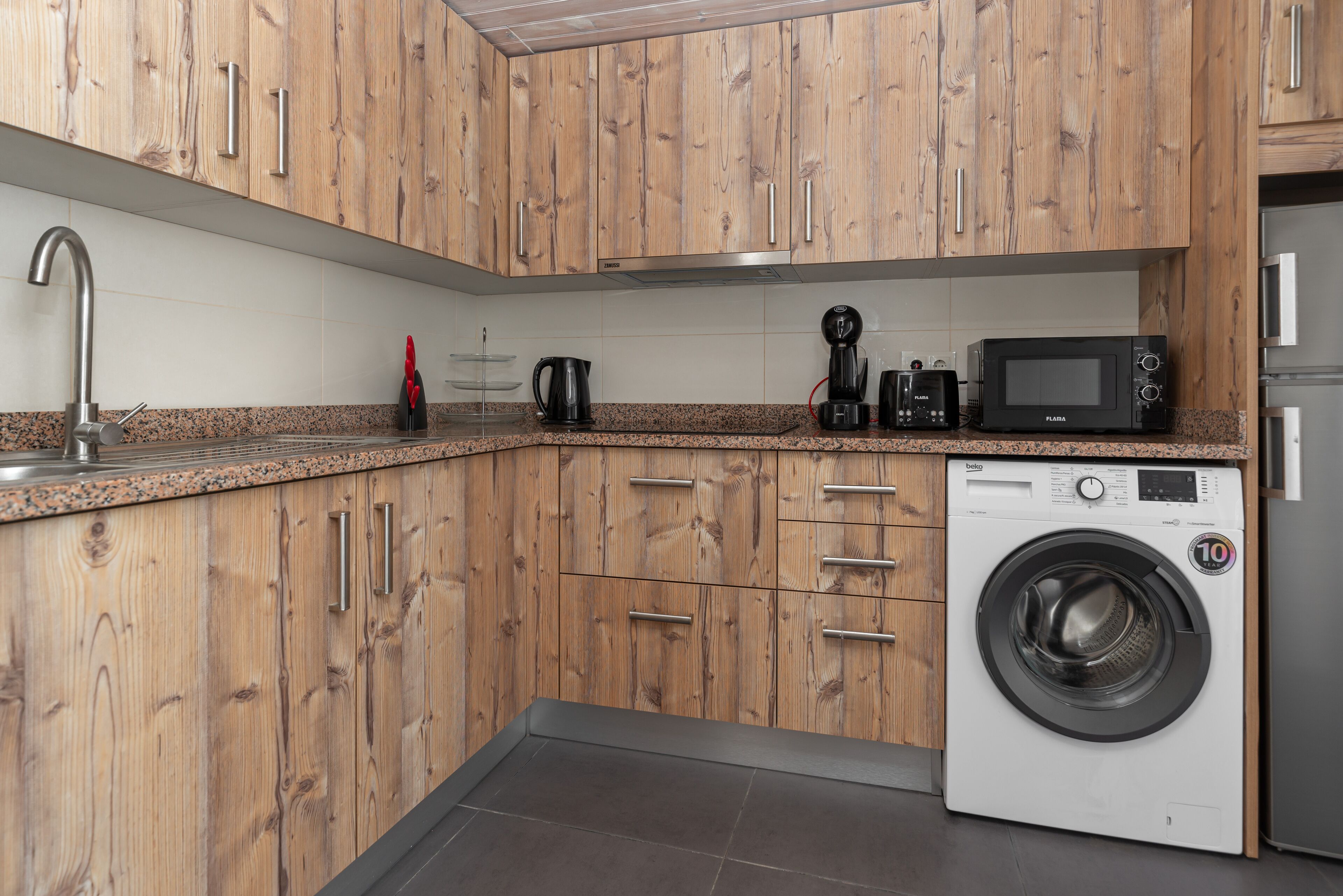 Una cocina moderna con armarios de madera, un lavamanos de acero inoxidable, una cafetera y una lavadora de carga frontal.