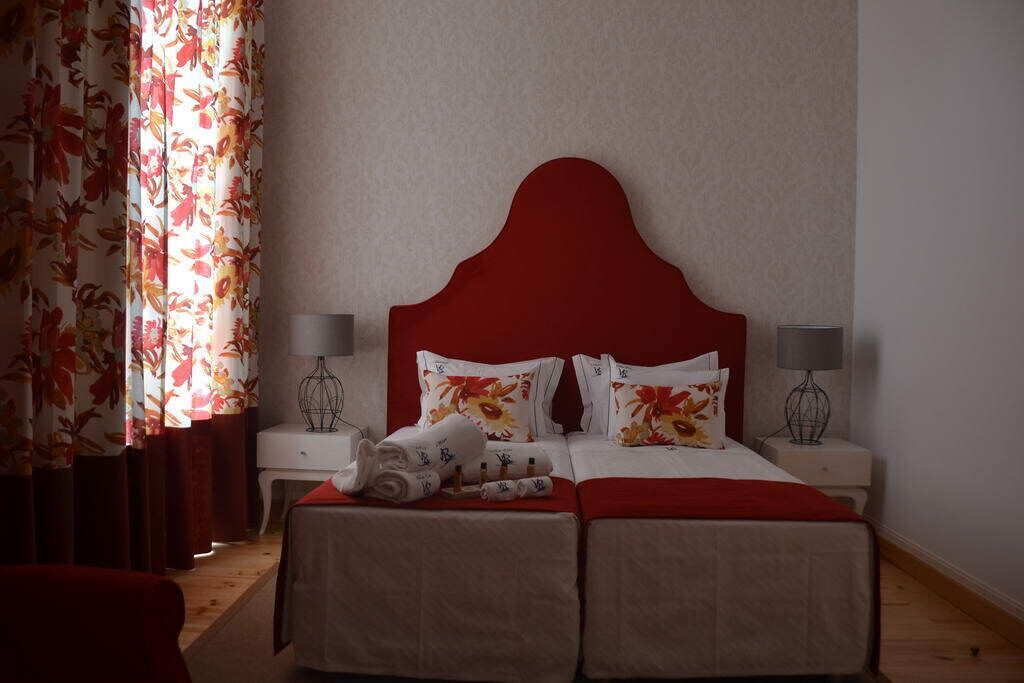 Habitación de hotel con cabecera roja, dos camas, cortinas florales, lámparas de noche y toallas dobladas con cuidado sobre las camas.