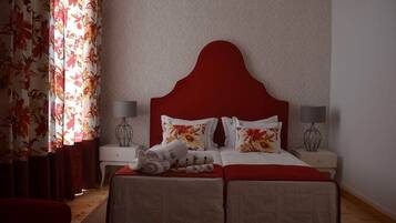 Habitación de hotel con cabecera roja, dos camas, cortinas florales, lámparas de noche y toallas dobladas con cuidado sobre las camas.