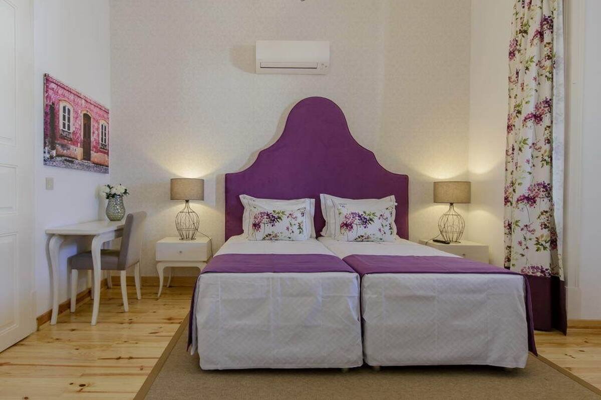 Ein Doppelbett mit violettem Kopfteil, zwei Nachttische mit Lampen, ein Schreibtisch mit Stuhl und ein blumiger Vorhang.