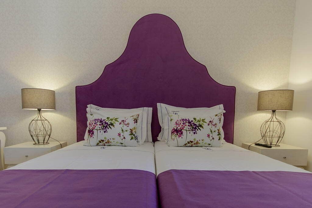 A double bed with a purple headboard, two bedside tables with lamps, and floral-patterned pillows.