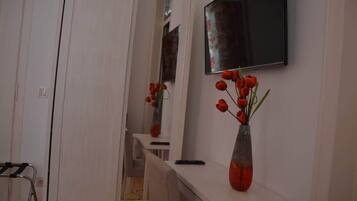 Een kamer met een witte kast, een spiegel, een aan de muur bevestigde televisie en een vaas met rode bloemen op een klein tafeltje.