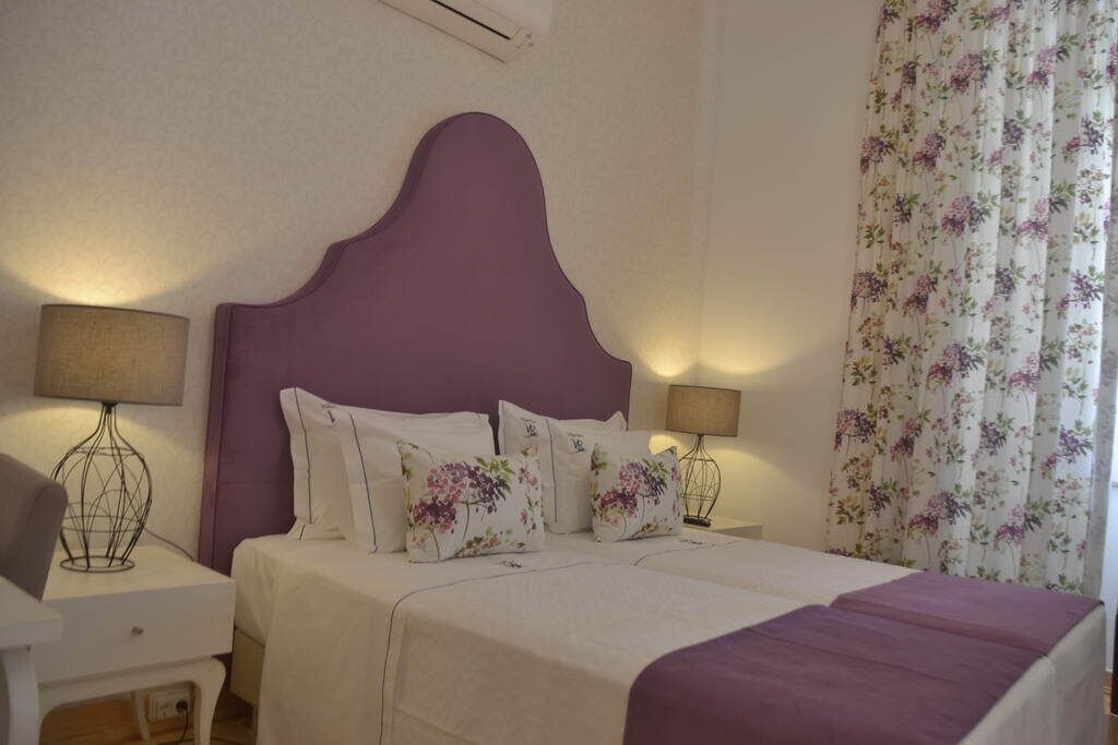 Una camera da letto con una testiera viola, due comodini con lampade, tende floreali e un letto con biancheria da letto bianca e cuscini a motivi floreali.