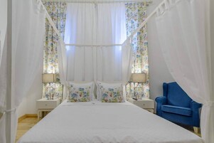A bedroom with a large bed, two bedside tables with lamps, a blue chair, and floral curtains.