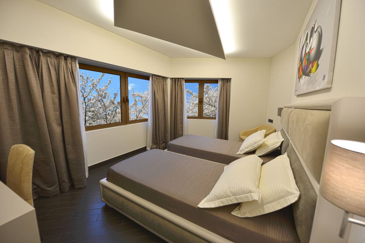 Une chambre d’hôtel avec deux lits, une chaise, une lampe et de grandes fenêtres avec des rideaux.