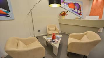 Un área de espera moderna con dos sillones color beige, una mesa de centro con revistas, un jarrón con flores y un cuadro abstracto de colores en la pared.