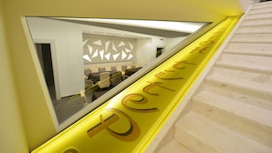 Una escalera con barandilla amarilla y un muro espejado que refleja una habitación con diseños geométricos en las paredes y una zona de estar.