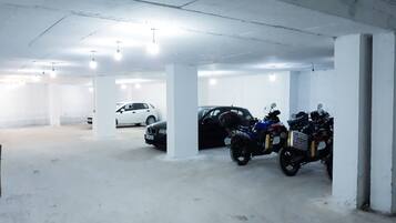 Un parking avec des piliers en béton, une voiture blanche, une voiture noire et plusieurs motos.