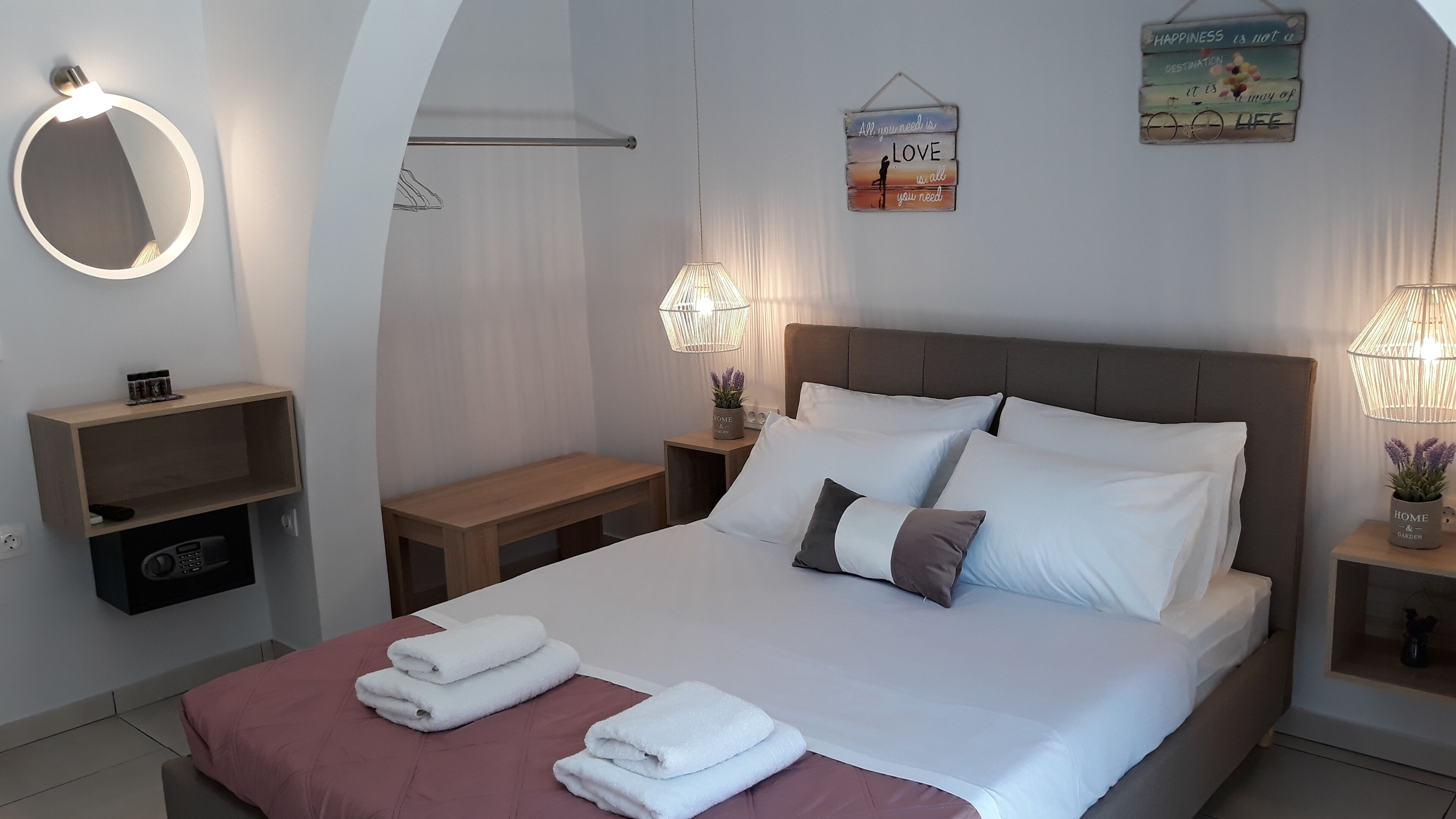 A neatly made bed with white linens and two pillows, a bedside table with a lamp, a wall-mounted mirror, and a small shelf with a few items.