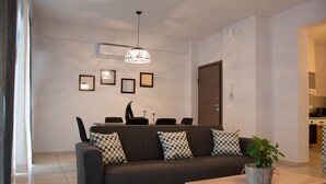 A modern living room with a sofa, coffee table, and decorative pillows. There is a dining area with chairs and a table, and a kitchen in the background. - B&M House (Kissamos)