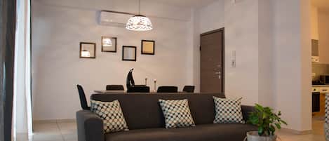 A modern living room with a sofa, coffee table, and decorative pillows. There is a dining area with chairs and a table, and a kitchen in the background.